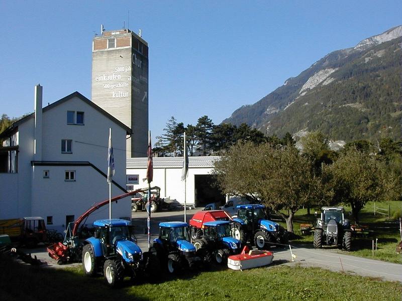 Das Betriebsareal an der Rheinmühle in Chur mit Wohnhaus, Silo-Turm, Halle und einer Reihe bereitgestellter Traktoren vor der Bergkulisse