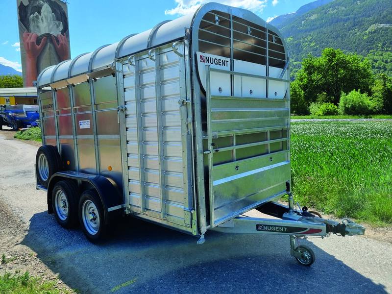 Verzinkter Nugent-Viehanhänger mit zwei Achsen auf einer Strasse, im Hintergrund Wiese und Berge